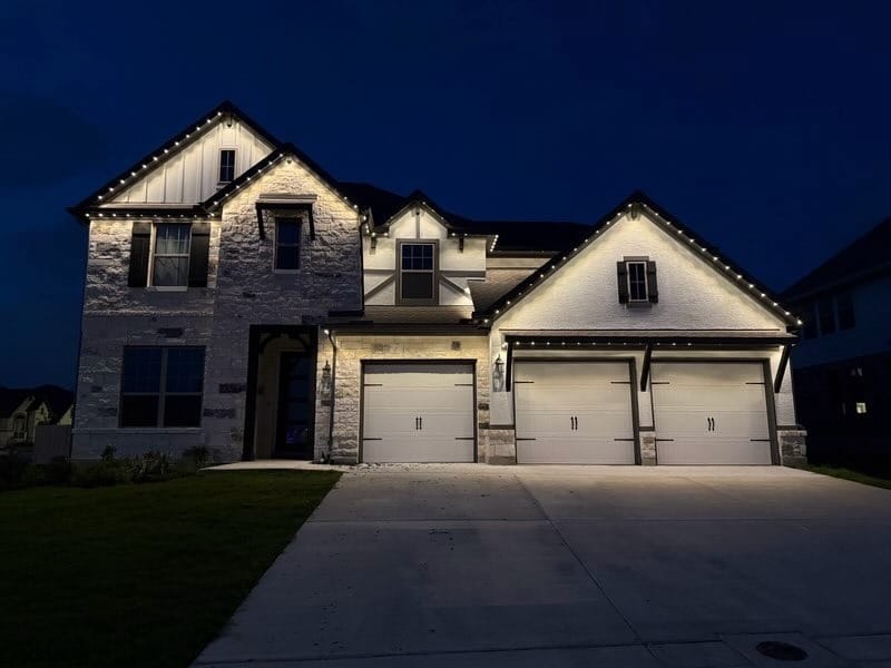 Real Dazzl install in Texas. Two-story stone home at night with permanent warm-white roofline lighting along the eaves and full accent uplighting illuminating the stone facade across all three garage bays and dormers.
